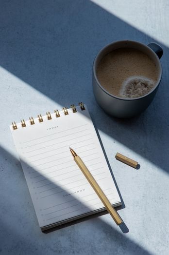 Image of a notepad, pencil and cup of coffee on a table. Image has blue tones.