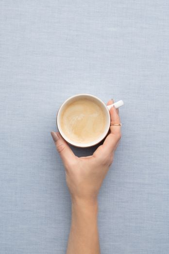 Photo of a hand grabbing a cup of coffee with creamer over a linen blue background.