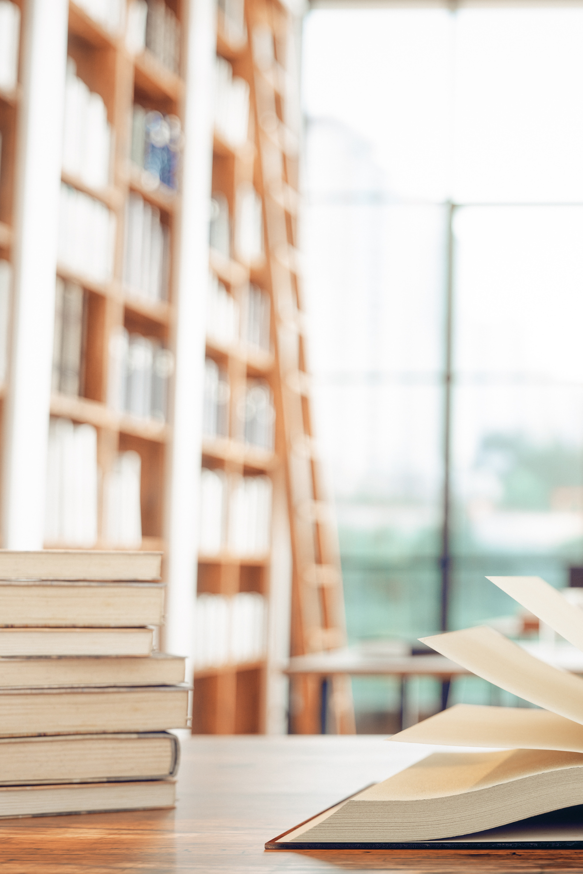 Stack of books and open book on table in bright sunny library.